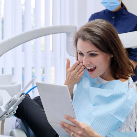 Happy middle aged woman with doctor dentist looking in mirror at teeth, sitting in dental chair. Medicine, dentistry and health care concept
