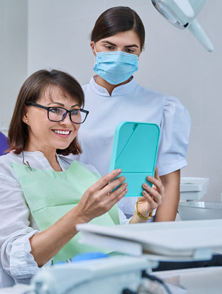 Happy middle aged woman together with dentist, patient sitting in dental chair looking at mirror. Prosthetics, treatment, implantation, dental teeth health, beauty care concept