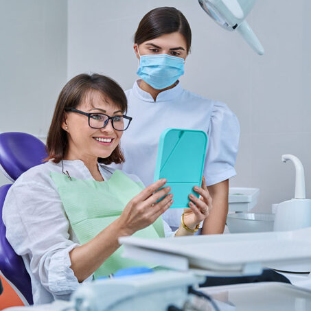 Happy middle aged woman together with dentist, patient sitting in dental chair looking at mirror. Prosthetics, treatment, implantation, dental teeth health, beauty care concept